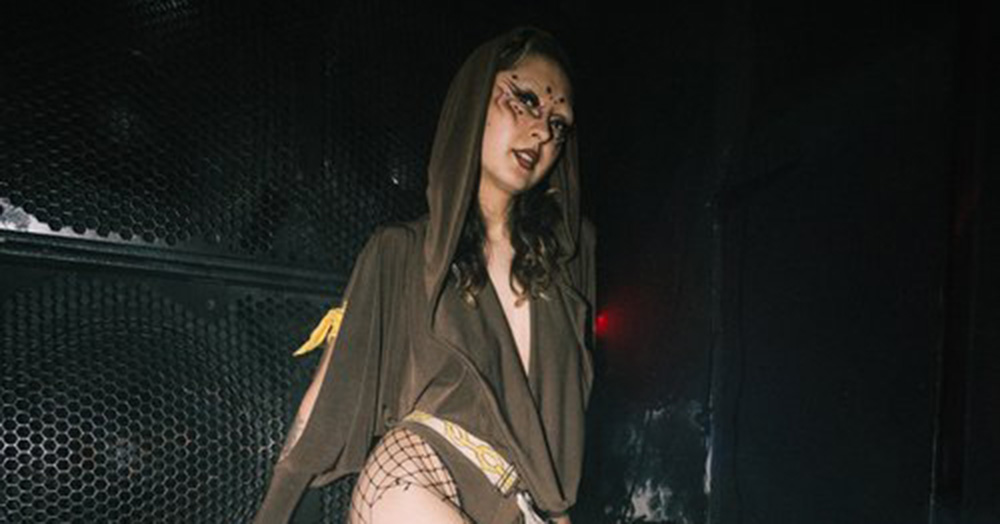 Image of Joyride Welfare Monitor, Tamaki. The person is photographed in a dark nightclub, wearing a hooded, low cut top, fishnet tights and bikini-like bottoms.