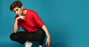 Photo of comedian Sarah Keyworth, crouching down against a blue backdrop, in a red t-shirt.