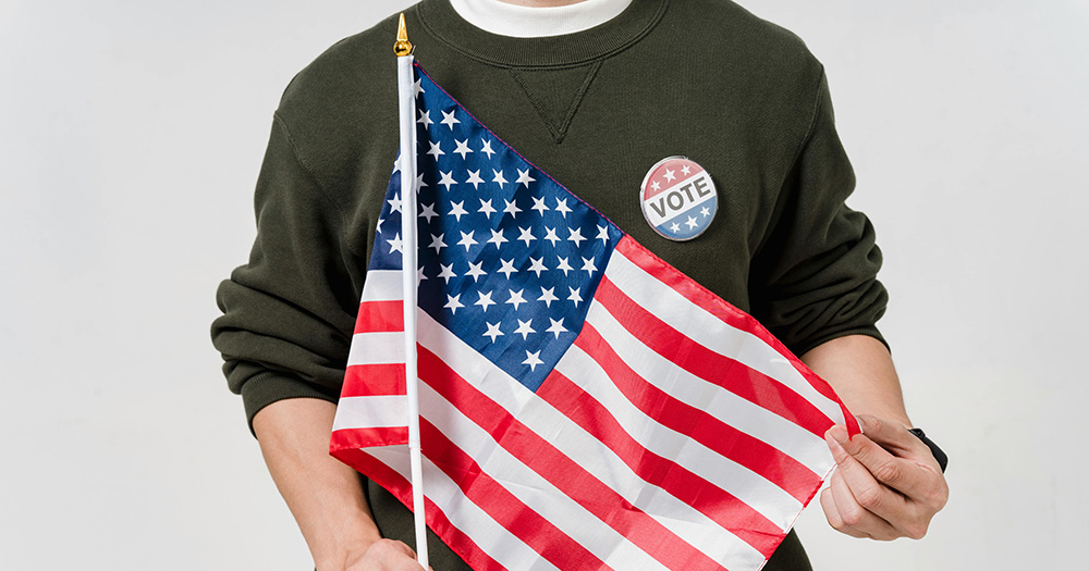 This article is about how trans people could be prevented from voting in the upcoming US elections. The image shows a person holding an American flag and wearing a 'vote' badge.