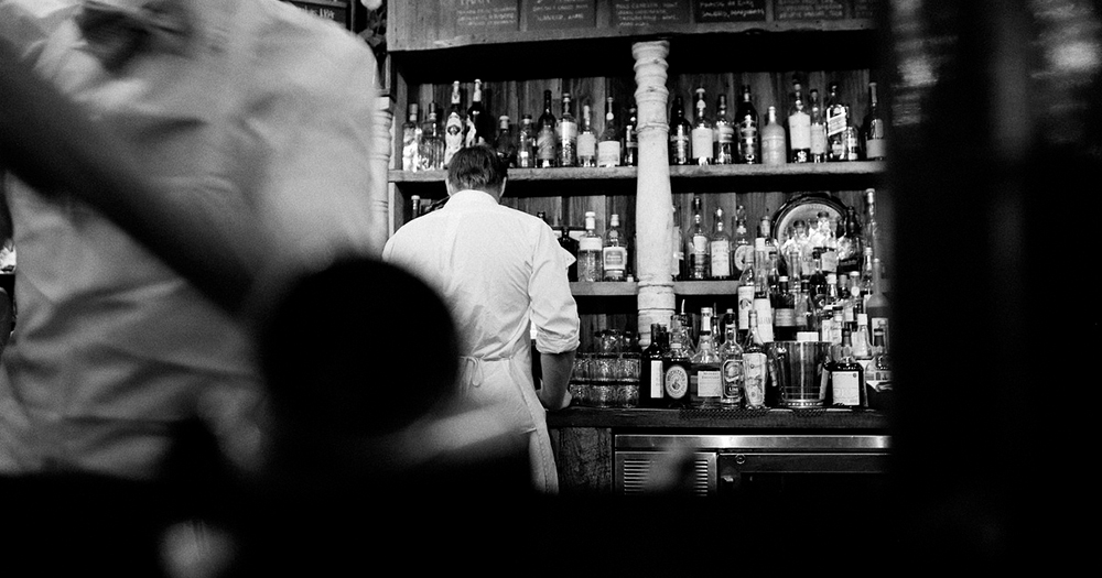 A black and white image of a barternder facing backwards from the counter while preparing a drink in a queer bar