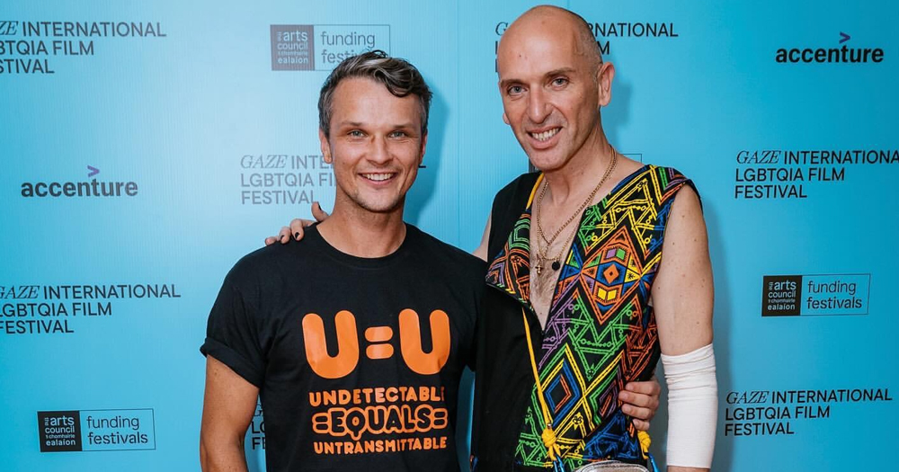 Poz Vibe hosts Robbie Lawlor and Veda Lady, who are hosting a fundraising event with GCN, posing on at an event with a blue background.