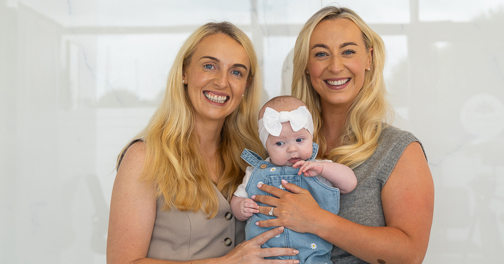 Lesley and Ger, a lesbian couple who became parents through IVF. In the photo, they are holding their baby and smiling at the camera.