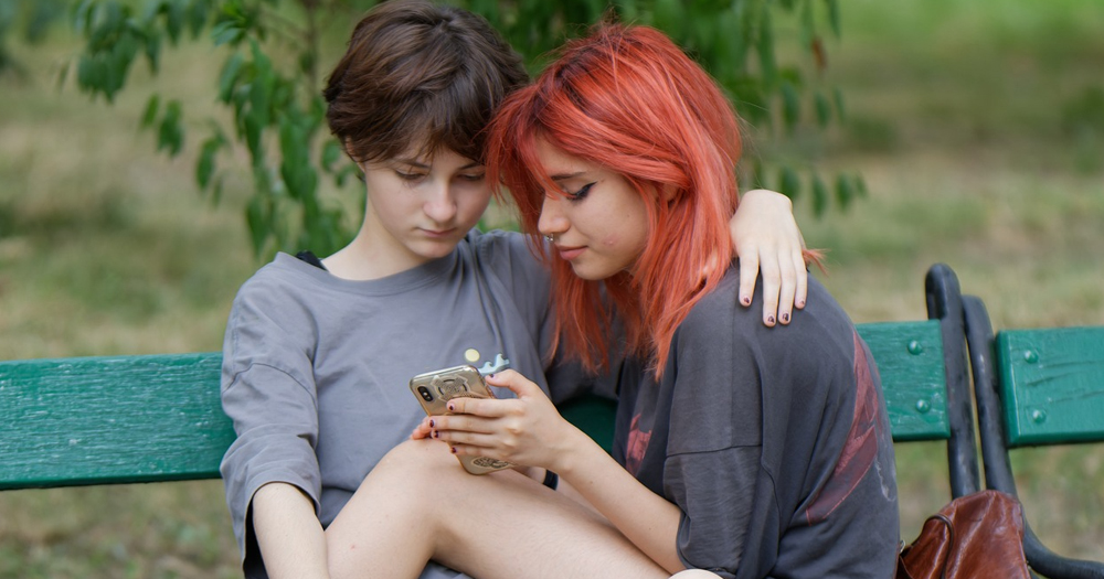 This article is about misogyny in the LGBTQ+ community. Two women sitting on a bench looking at a phone screen