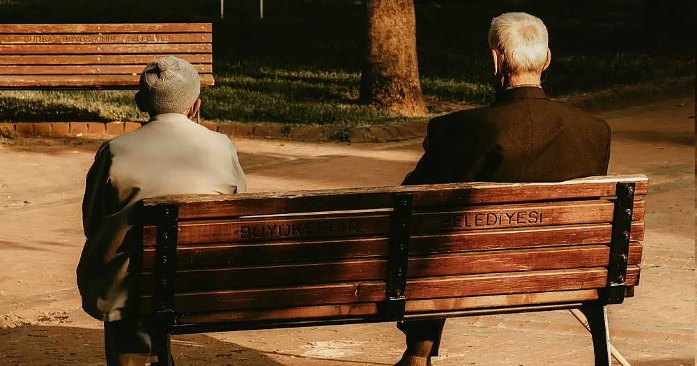 This article is about older queer people coming out later in life. Two older men sitting on a bench in a park, with their back facing the camera.