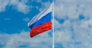 This article is about Russia advancing a bill to ban adoptions by people from countries where gender-affirming care is legal. in the photo, a Russian flag flying with a blue sky in the background.