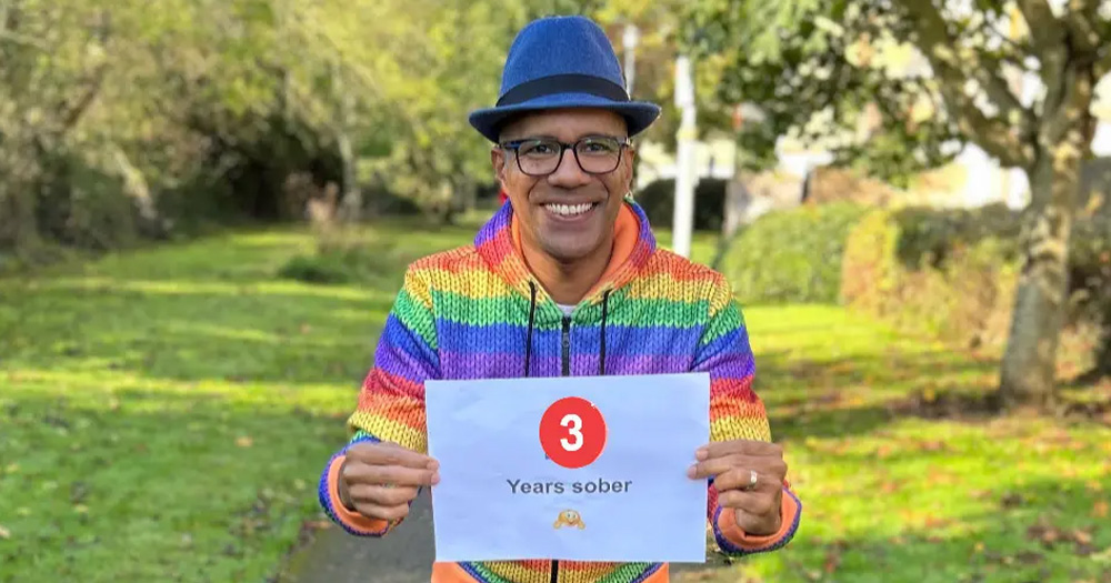 An image of Marlon Jimenez-Compton celebrating three years of sobriety. He wears a rainbow jumper, a fedora and holds a sign reading "3 years sober".