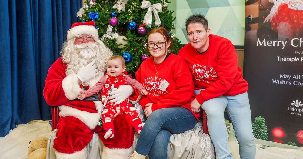 Photo of two lesbian mothers posing with their baby and Santa Claus, all wearing bright red clothes for Christmas.