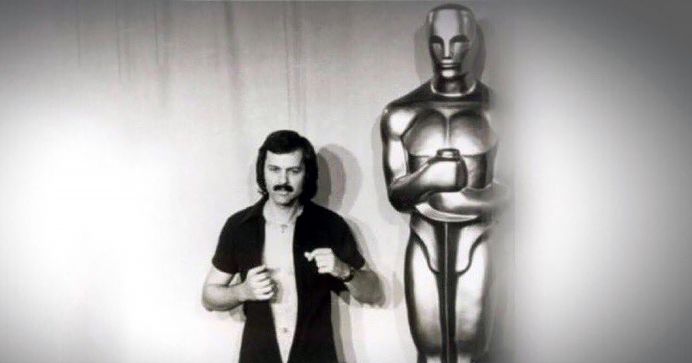 Black and white photo of Robert Opel standing next to the Academy Awards statue while undressing.