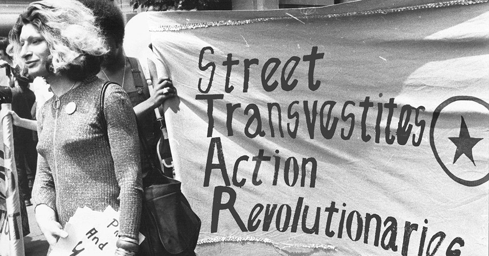 Image of Sylvia Rivera marching beside a banner reading Street Transvestites Action Revolutionaries.