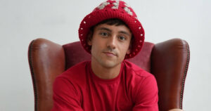 Tom Daley, who has just announced a new TV show, sitting on a chair wearing a red t-shirt and knitted hat.