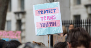 This article is about the UK's puberty blockers ban. The image shows a protest sign reading "protect trans youth".