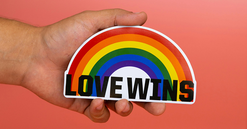 This article is about inclusive brand advertising. The image shows a had holding a small rainbow cut-out graphic that reads "Love Wins".