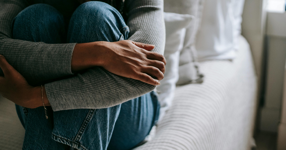 This article is about intimate partner violence in LGBTQ+ relationships. In the photo, a person sitting on a bed hugging their knees.