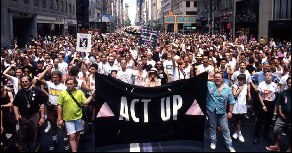 This article is about LGBTQ+ liberation in the US. The image shows an ACT UP New York protest, with large crowds of people walking through the streets of Manhattan.