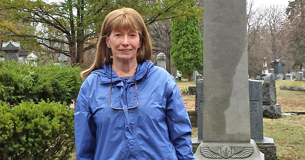 Prof. Lynn Conway standing in a graveyard wearing a blue rain jacket.