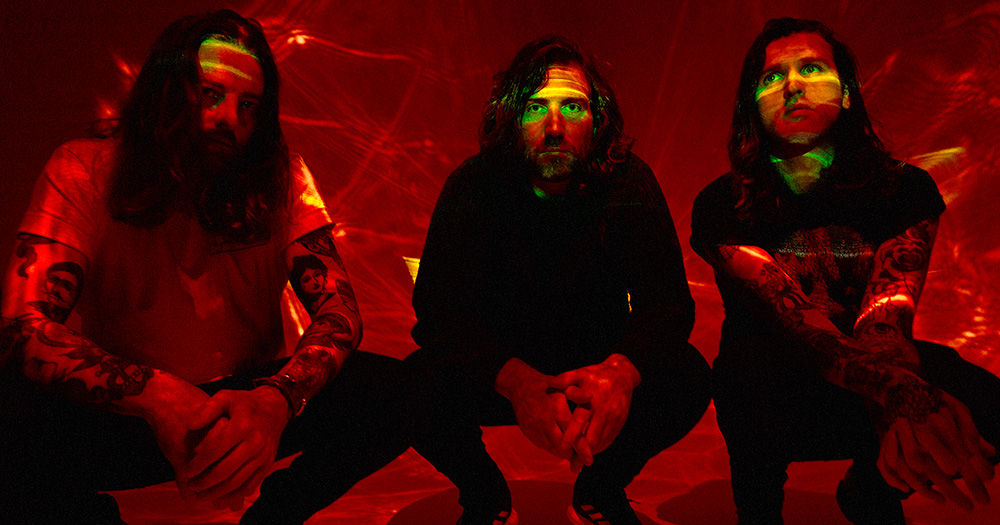 A press image of TAYNE. The trio is pictured crouching down in red lights.