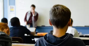 School children sit in a classroom while a teacher is at a whiteboard.