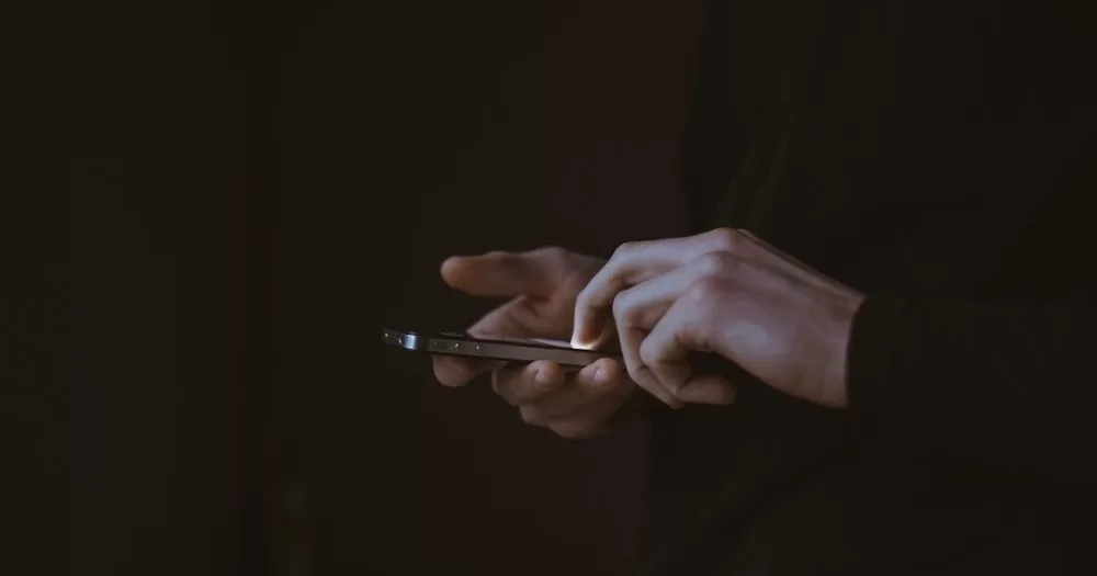 This article is about how to stay safe from sextortion on Grindr. In the photo, the hands of a person holding a phone in the dark.
