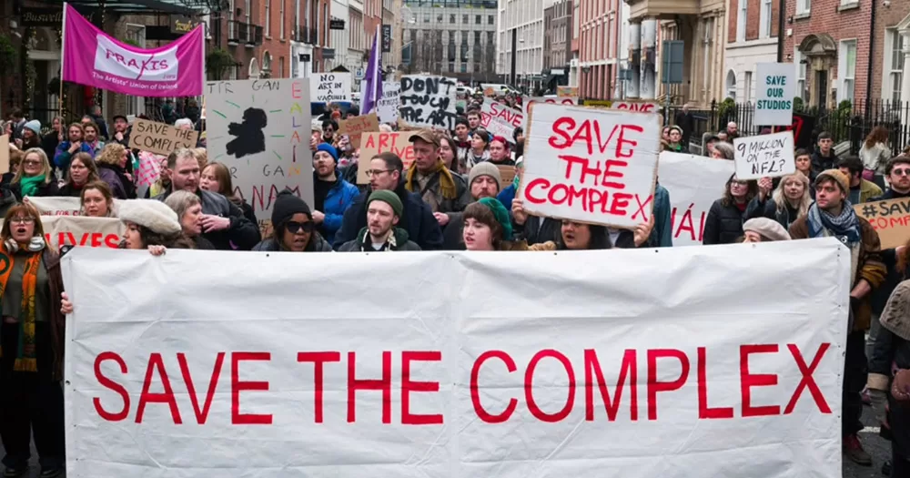 People protesting to save The Complex after the arts centre announced its closure. Marching in the streets, people are holding banners that read 'Save the Complex'.