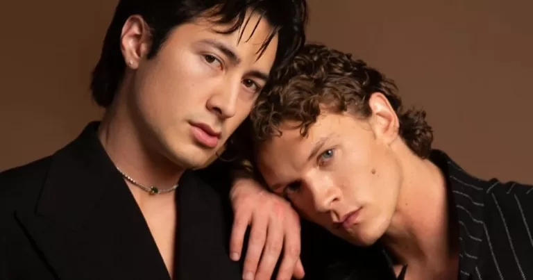 The image shows Heated Rivalry stars Hudson Williams and Connor Storrie. Hudson is on the left, wearing a black suit and a small necklace. Connor is leaning his head on Hudson's shoulder, also dressed in black, with his arm resting on Hudson's shoulder. Both are staring at the camera. Season 2 filming will begin this August.