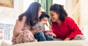 This article is about same-sex mothers in Italy. In the photo, two women holding a child on a bed and smiling.