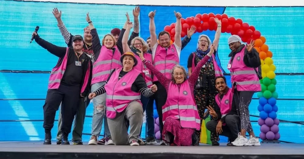 Photo of the Midlands LGBT+ Project team, who are looking for volunteers, on a stage wearing bright pink vests and posing for a group photo.