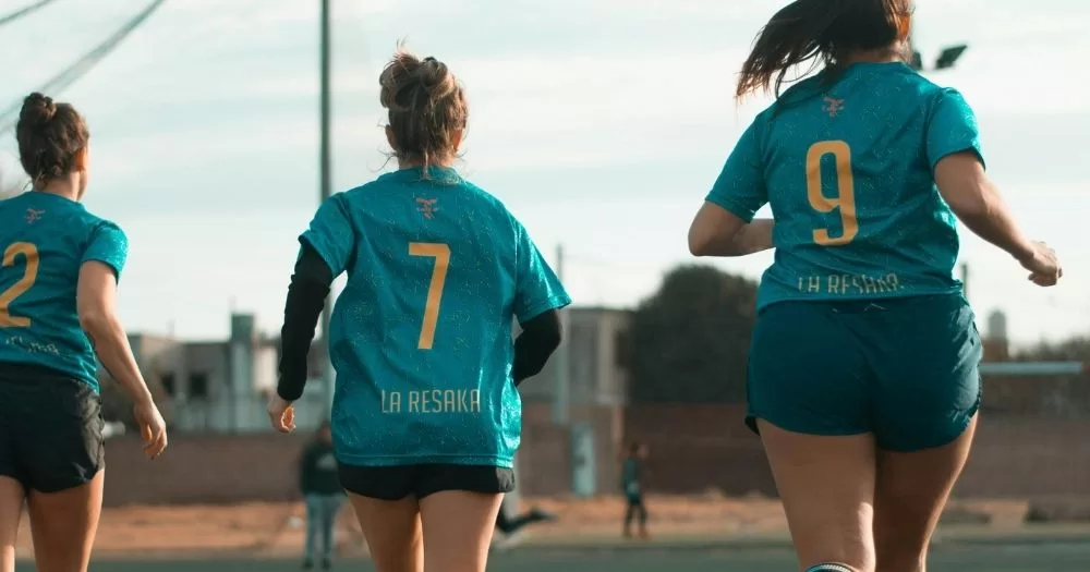 A trio of women running together on a sports field. They are in a sports uniform.