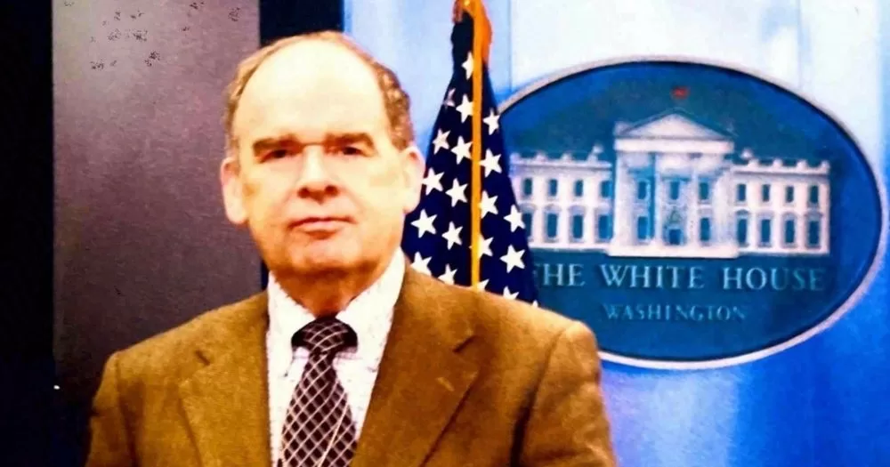 Sean J. Connolly stands in front of a US flag and a White House crest.