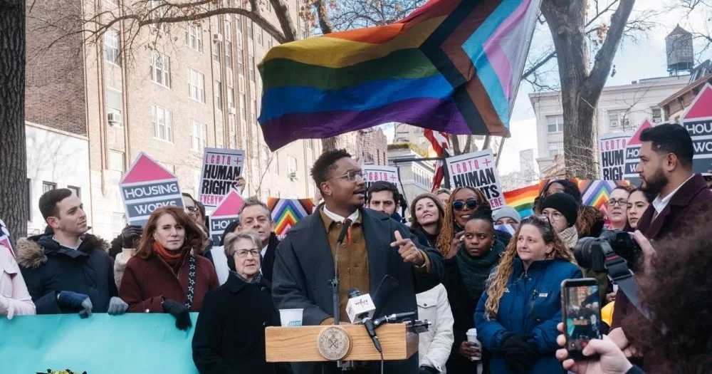 The image shows Council Member Chi Ossé standing at a platform at the Stonewall Monument and speaking, with a large pride flag in the background and a crowd gathered holding flags and placards.