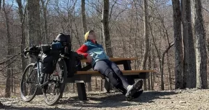 Dad cycles America for Trans American Ride 2026. Ray sits on a park bench, next to a bicycle and among some trees.