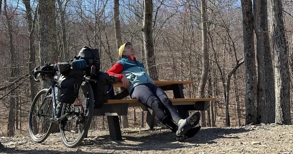 Dad cycles America for Trans American Ride 2026. Ray sits on a park bench, next to a bicycle and among some trees.